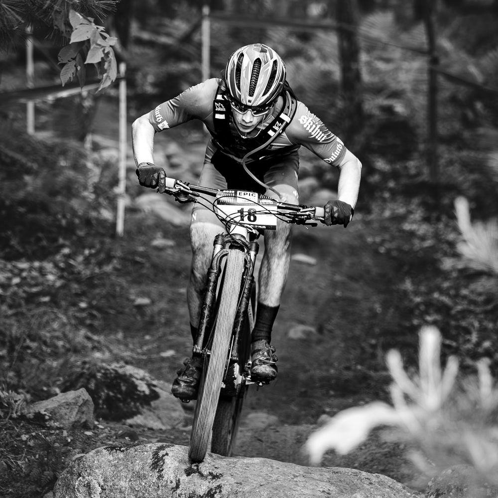 A rider dropping a front wheel off the edge of a large rock slab, committed and looking down the trail.