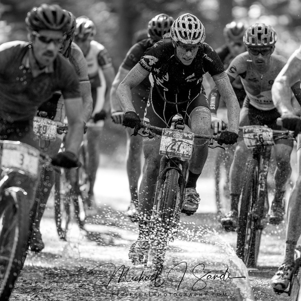 A pack of racers riding hard through a flooded section of wooded singletrack, spray coming off every wheel.
